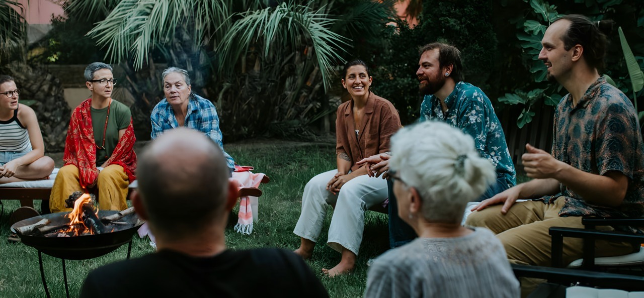 A diverse group of people sitting in a circle outdoors around a small fire pit, engaged in conversation and smiling.