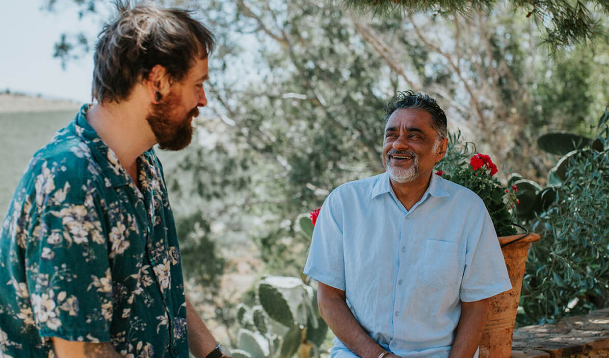 Two men smiling and conversing outdoors in a garden with potted plants and cactus in the background.