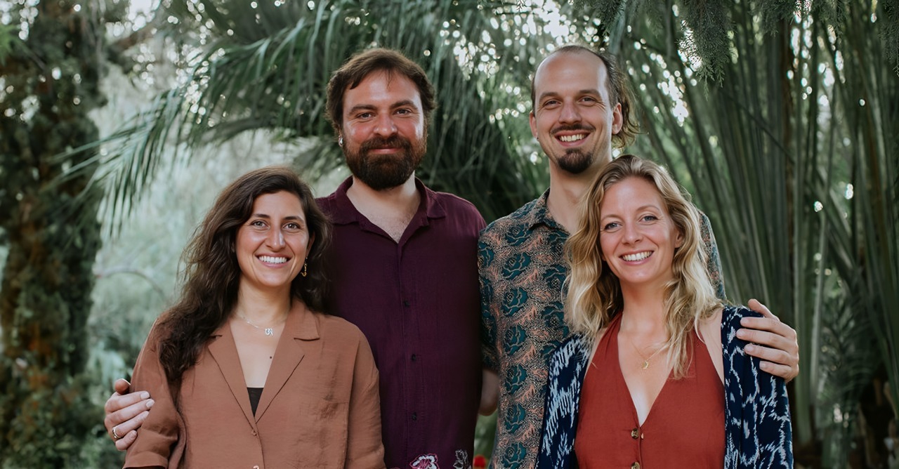 Group of two men and two women smiling and standing closely together outdoors with greenery in the background.