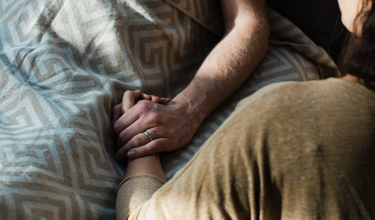 Two people holding hands, one lying in bed under a patterned blanket, conveying support and comfort.