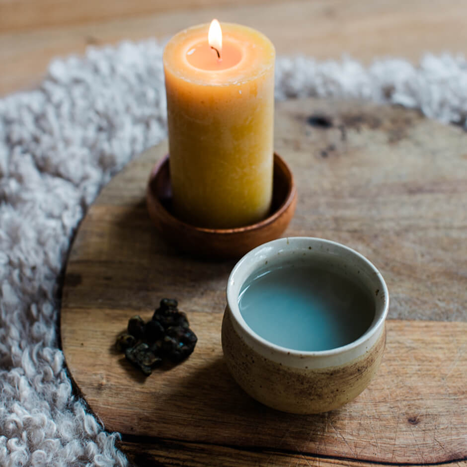 Lit yellow candle, small ceramic cup with liquid, and a few dark dried herbs on a wooden board.