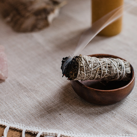 Lit sage smudge stick emitting smoke resting in a small wooden bowl on a textured beige cloth.