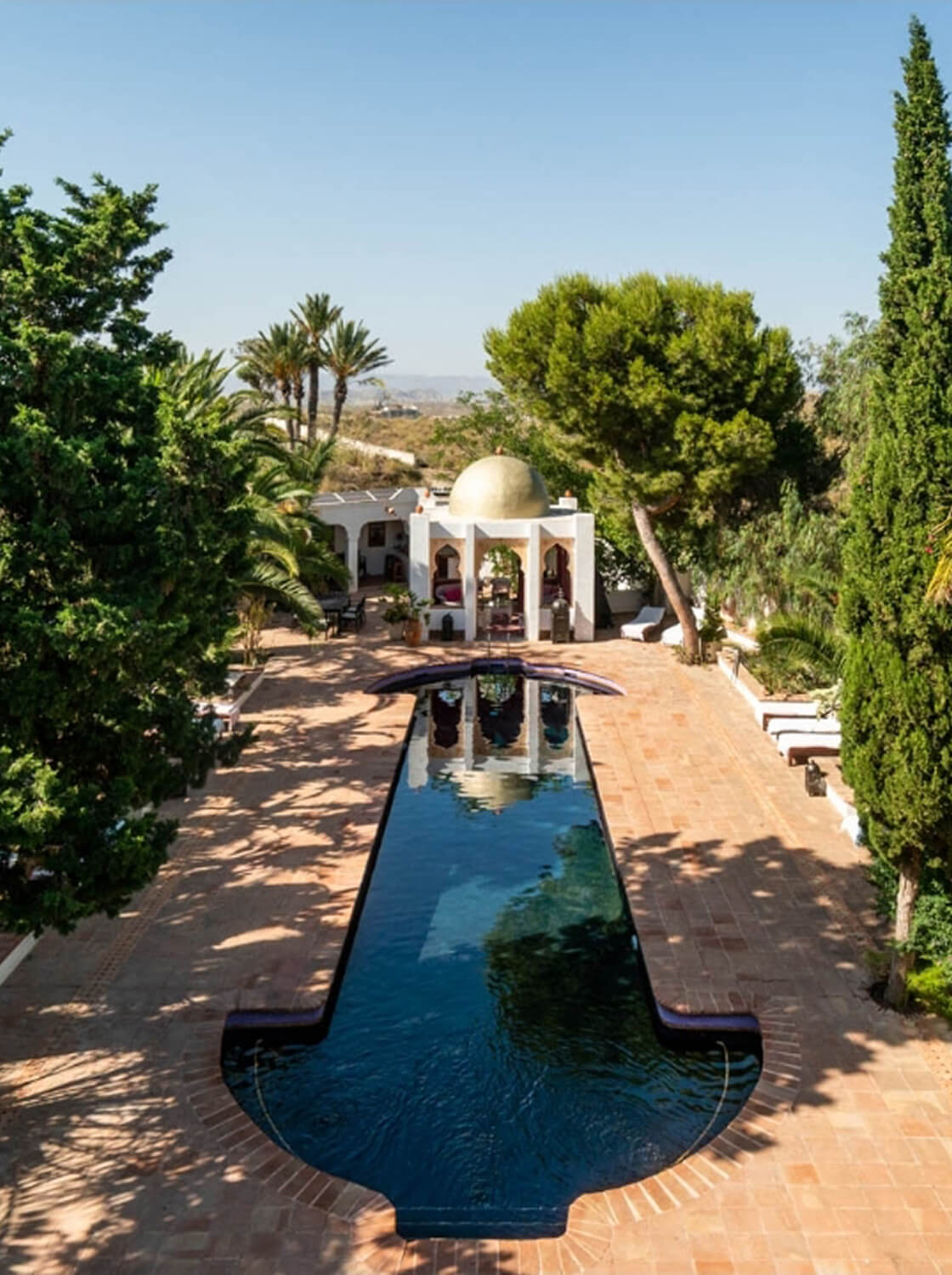 Long rectangular pool with rounded ends surrounded by terracotta tiles, trees, and a white gazebo with a golden dome in the background under a clear blue sky.