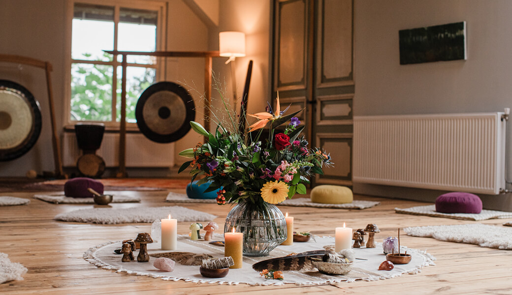 A serene room with a floral centerpiece surrounded by lit candles, crystals, incense, and cushions on a wooden floor.