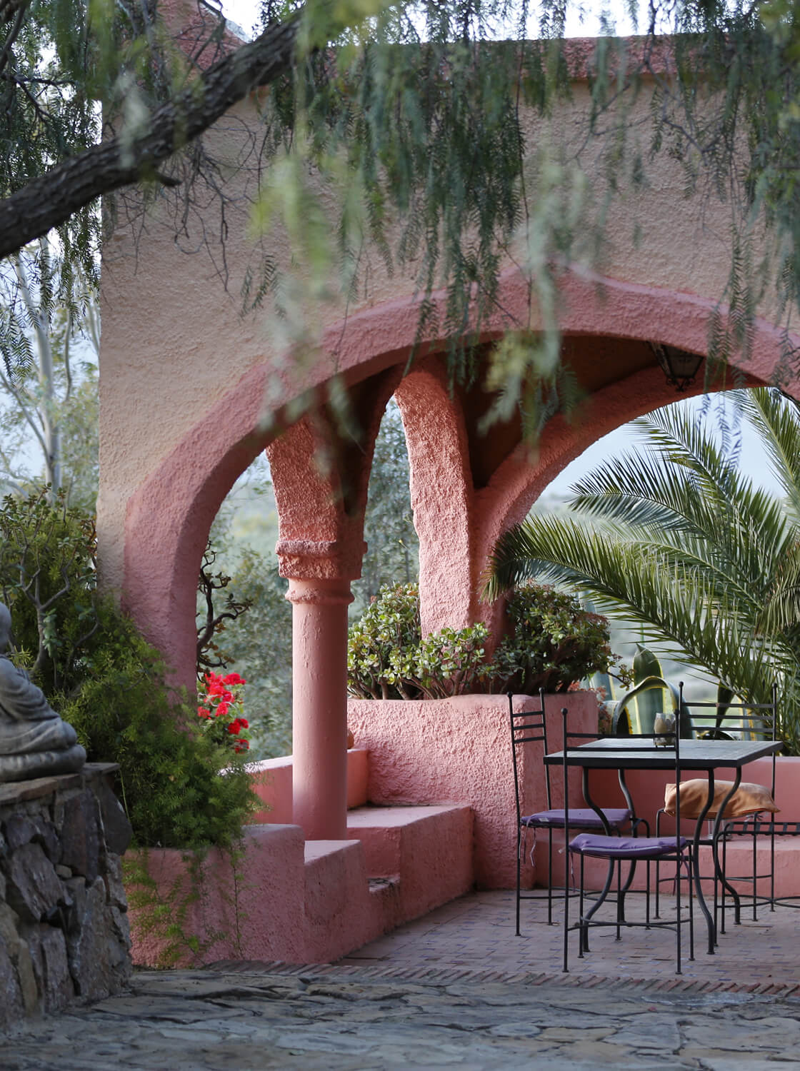 Outdoor patio with pink stucco arches, black metal table and chairs with purple cushions, surrounded by green plants and trees.