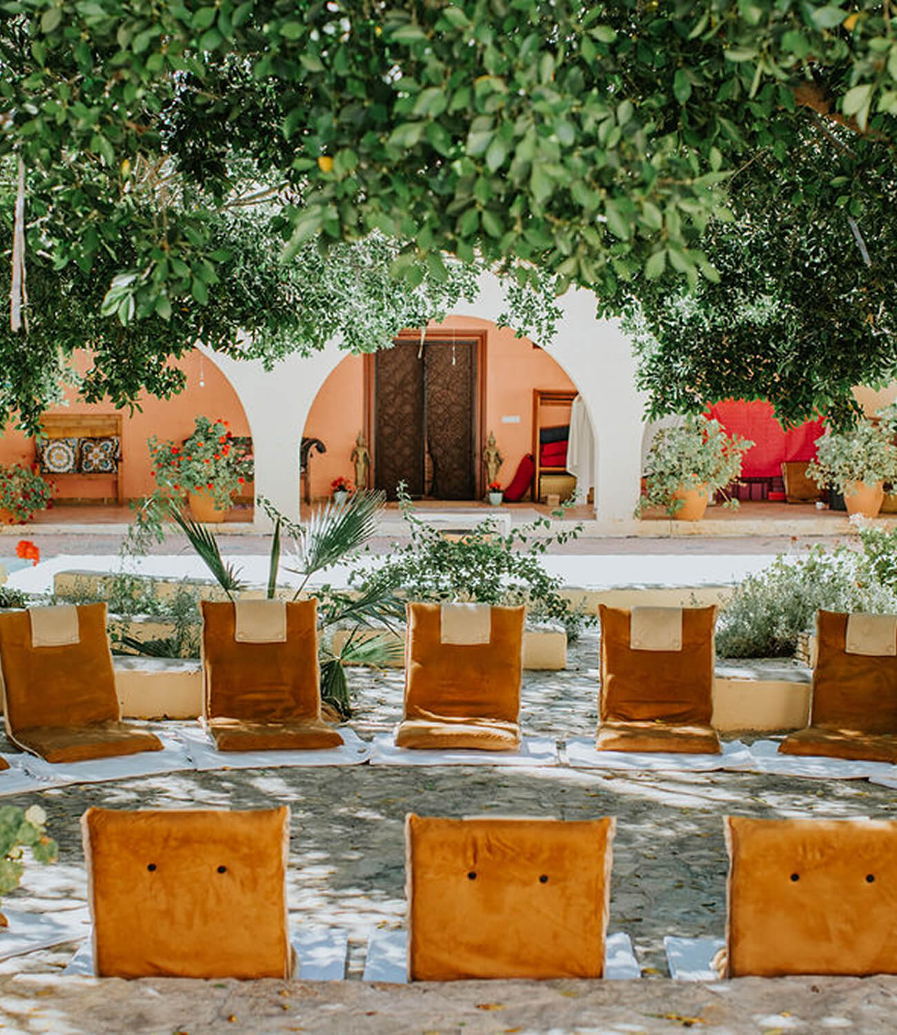 Outdoor seating area with cushioned brown chairs arranged in a circle under lush green trees, in front of a building with white arches and potted plants.