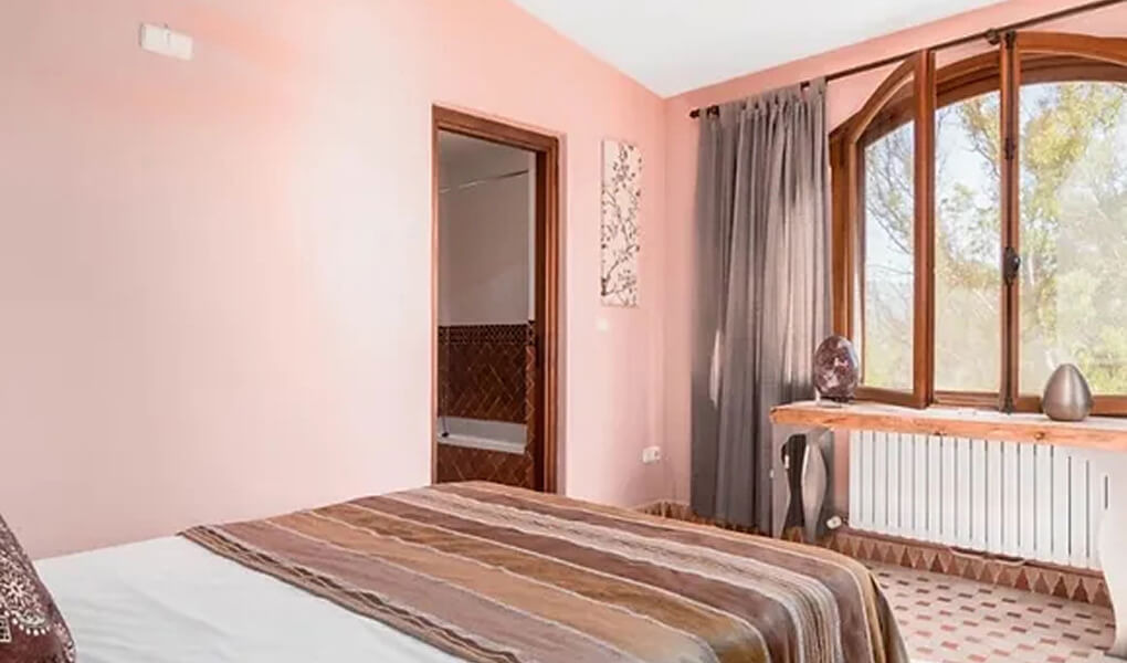 Bedroom with a large bed covered in a brown striped blanket, a pink wall, an open door revealing a bathroom, and a curtained window overlooking trees.