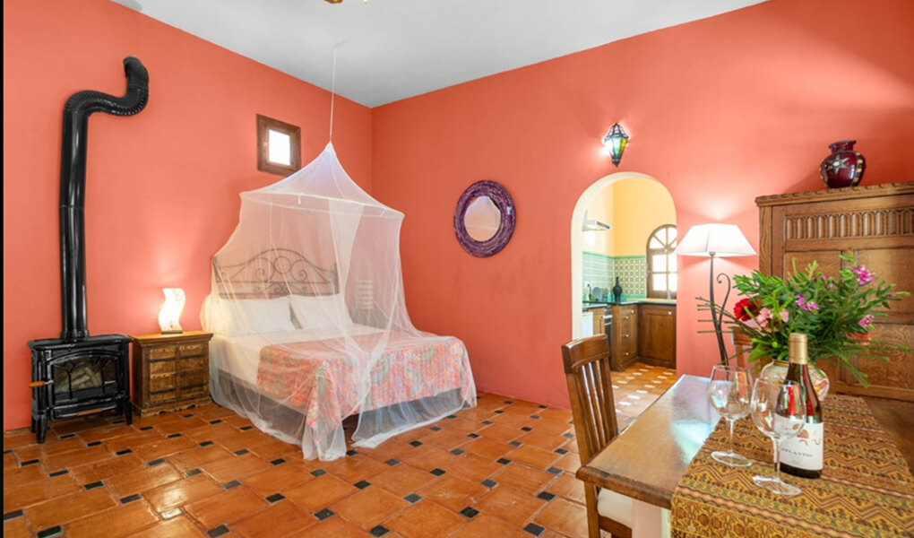 Cozy bedroom with a canopy-covered bed, wood stove, orange walls, and a wooden table set with wine and flowers.