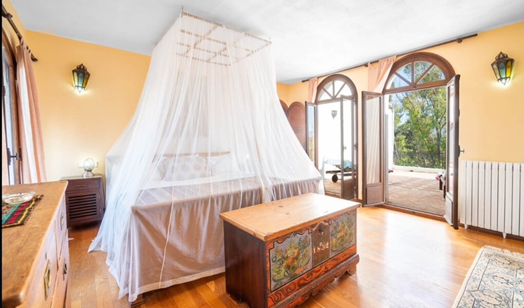 Bright bedroom with a canopy bed covered by white sheer curtains, wooden furniture, and arched doors opening to a sunny outdoor terrace with trees.