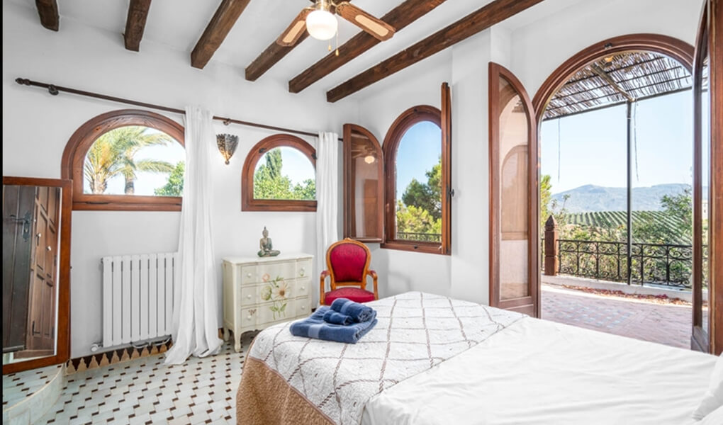 Bright bedroom with wooden ceiling beams, arched windows and doors opening to a terrace overlooking greenery and mountains.