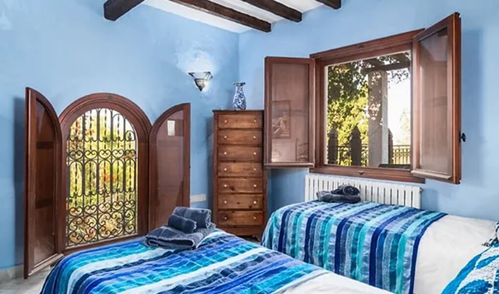 Room with two single beds dressed in blue striped bedding, wooden window and door open to garden with decorative grille, and wooden chest of drawers against light blue walls with exposed ceiling beams.