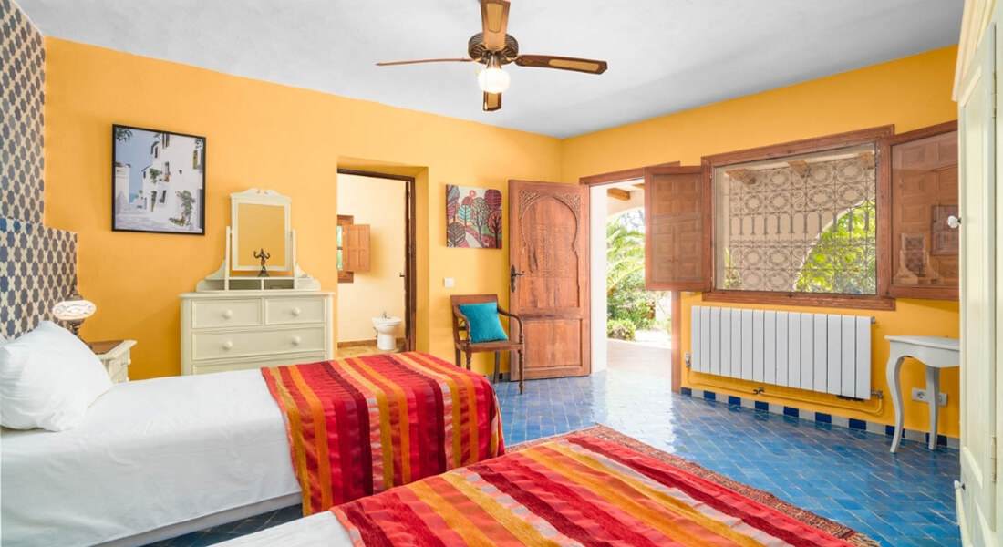 Bright room with two single beds featuring red and orange striped blankets, yellow walls, blue tiled floor, wooden door, and open window with patterned curtain.