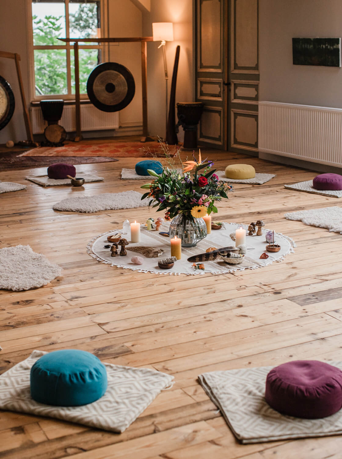 Cozy meditation room with circular arrangement of floor cushions and mats around a centerpiece of flowers, candles, and crystals on wooden floor.