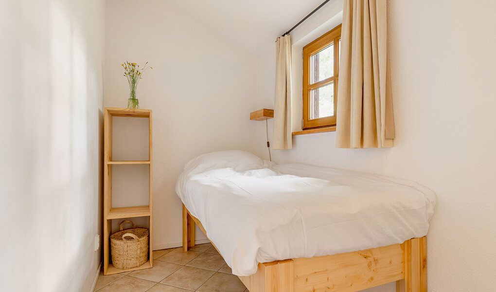 Small minimalist bedroom with single wooden bed, white bedding, wooden shelving unit with basket and flowers, and window with beige curtains.