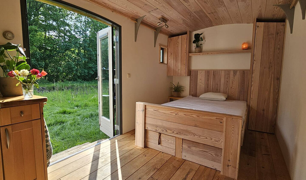 Interior of a small wooden bedroom with a bed, plants, and an open door leading to a green grassy area.
