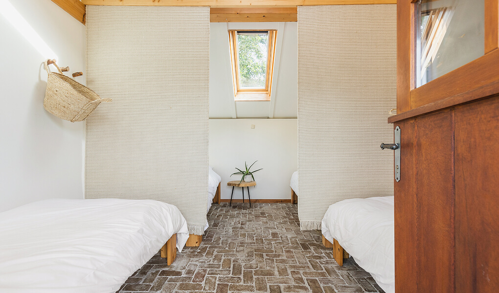 Minimalist shared bedroom with two single beds separated by beige curtains, a skylight window, and a small wooden stool with a potted plant in the center.