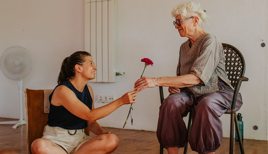 An elderly woman sitting on a chair hands a pink carnation flower to a younger woman seated on the floor in a cozy room.