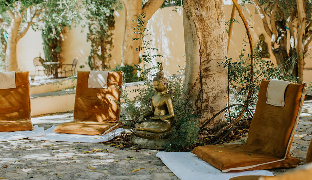 Outdoor meditation area with brown floor cushions arranged on white mats around a bronze Buddha statue, shaded by trees.