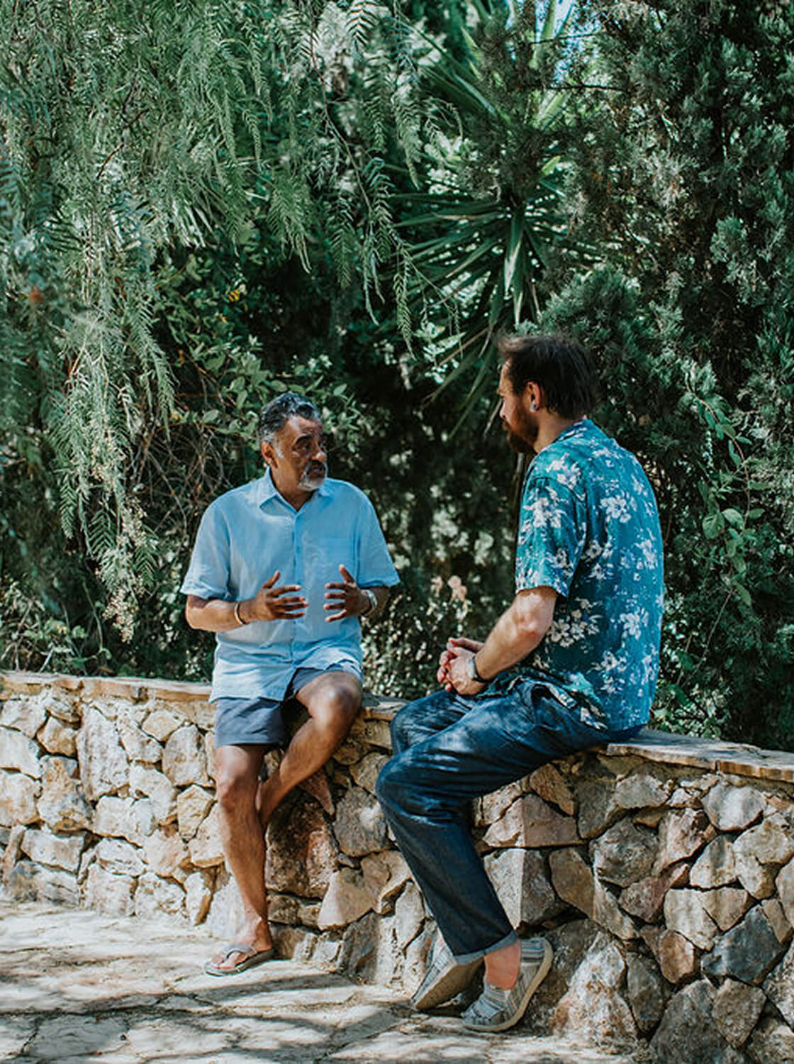Two men sitting on a stone wall outdoors, engaged in conversation surrounded by lush greenery.