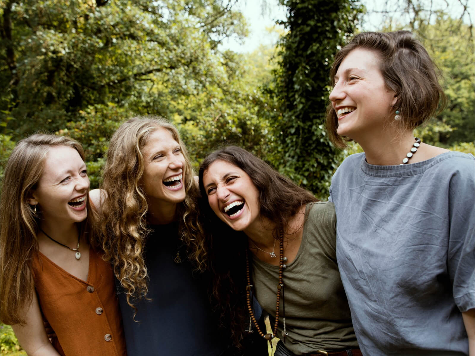 Four women standing closely outdoors, laughing joyfully together with a background of green trees.