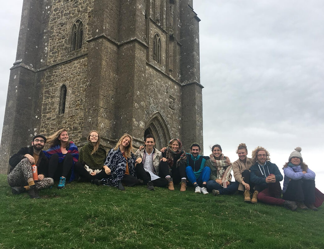 Group of eleven people sitting on grass in front of a large stone tower under a cloudy sky.