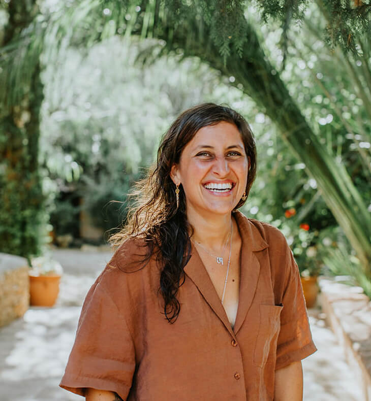 Smiling woman with wavy dark hair wearing a brown shirt, standing outdoors with trees and plants in the background.