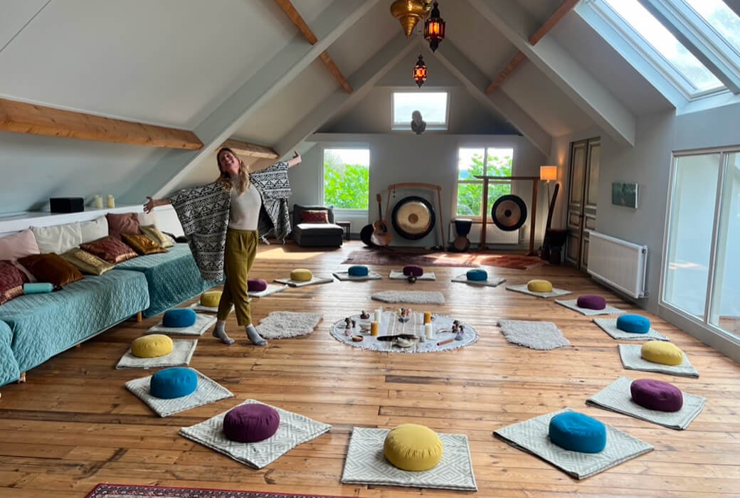 Woman standing with arms outstretched in a spacious attic room set up with colorful meditation cushions arranged in a circle on wooden floor.
