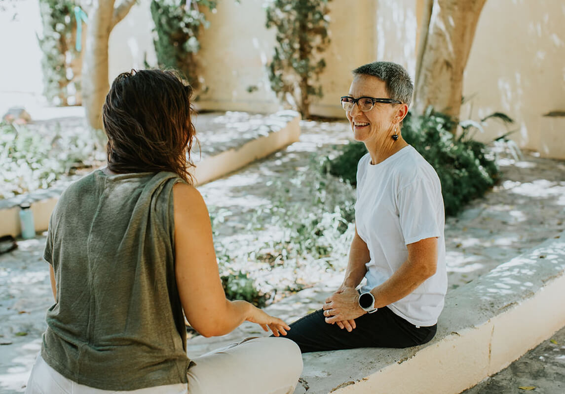 Two women sitting outdoors on a low stone wall, engaged in conversation, with greenery and sunlight in the background.