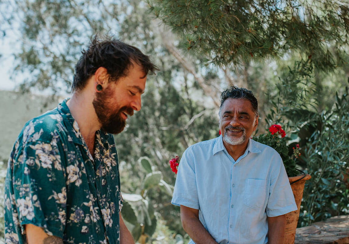 Two men smiling and enjoying a conversation outdoors surrounded by greenery and red flowers.