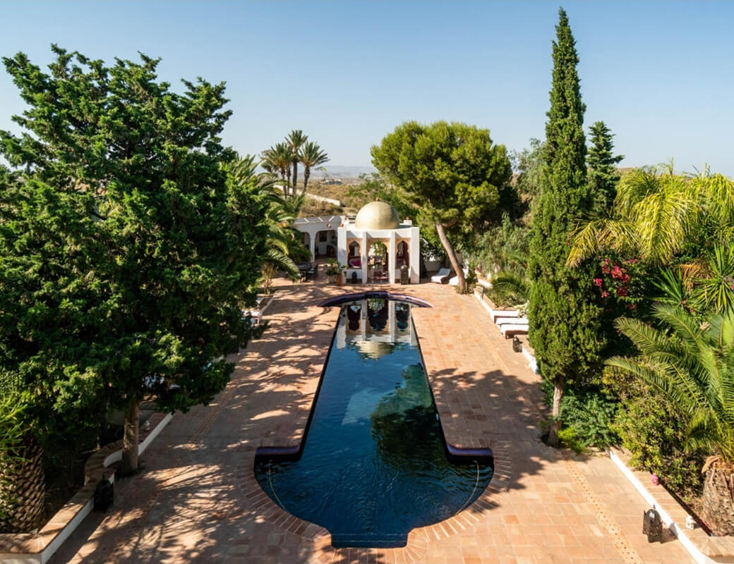 Outdoor pool with a unique shape surrounded by trees and a Mediterranean-style pavilion with a golden dome under a clear blue sky.