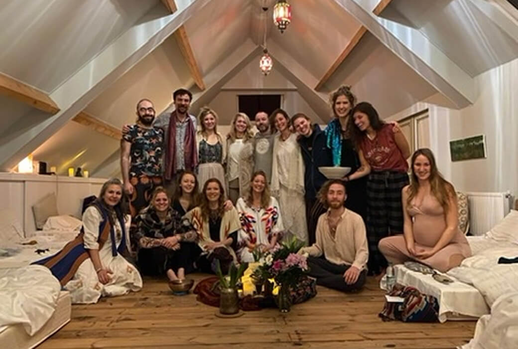 Group of 15 people gathered in a cozy attic room with wooden floor, some sitting and some standing close together, surrounded by plants, candles, and soft lighting.