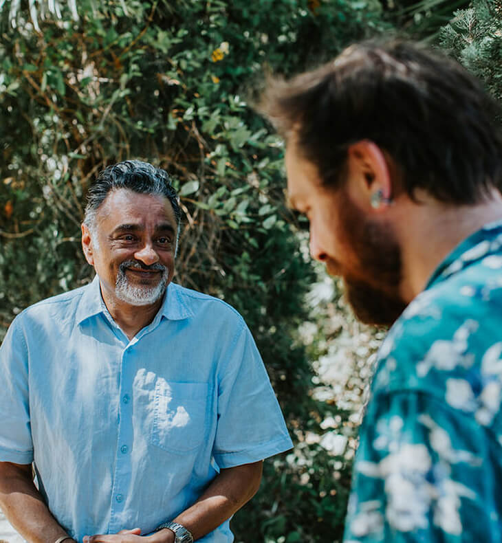 Two men smiling and engaging in conversation outdoors with green foliage in the background.