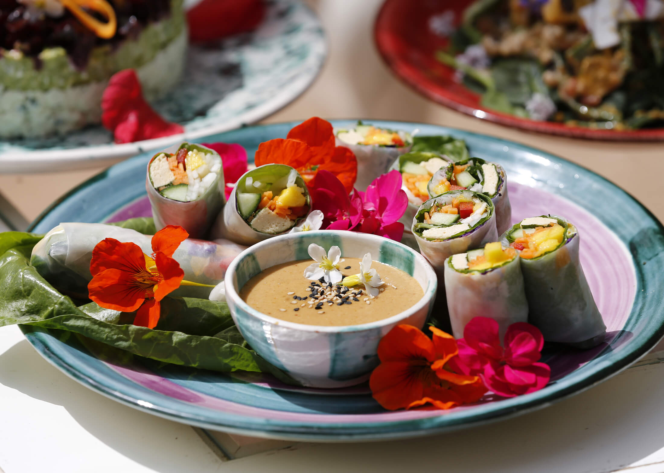 Plate with fresh vegetable spring rolls, colorful edible flowers, and a bowl of creamy dipping sauce topped with sesame seeds.