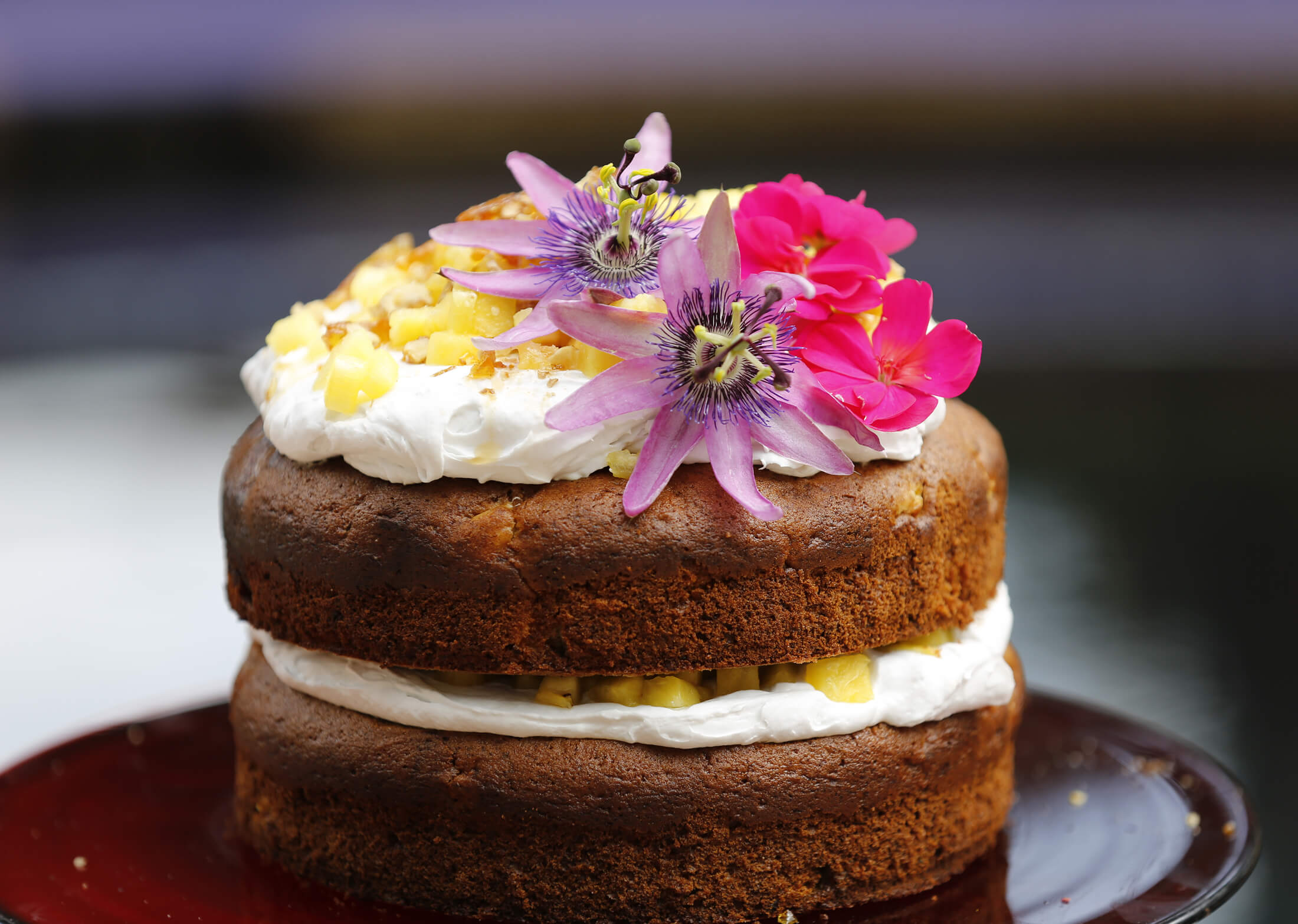 Two-layer cake with white cream and diced pineapple filling, topped with whipped cream and vibrant purple and pink edible flowers.