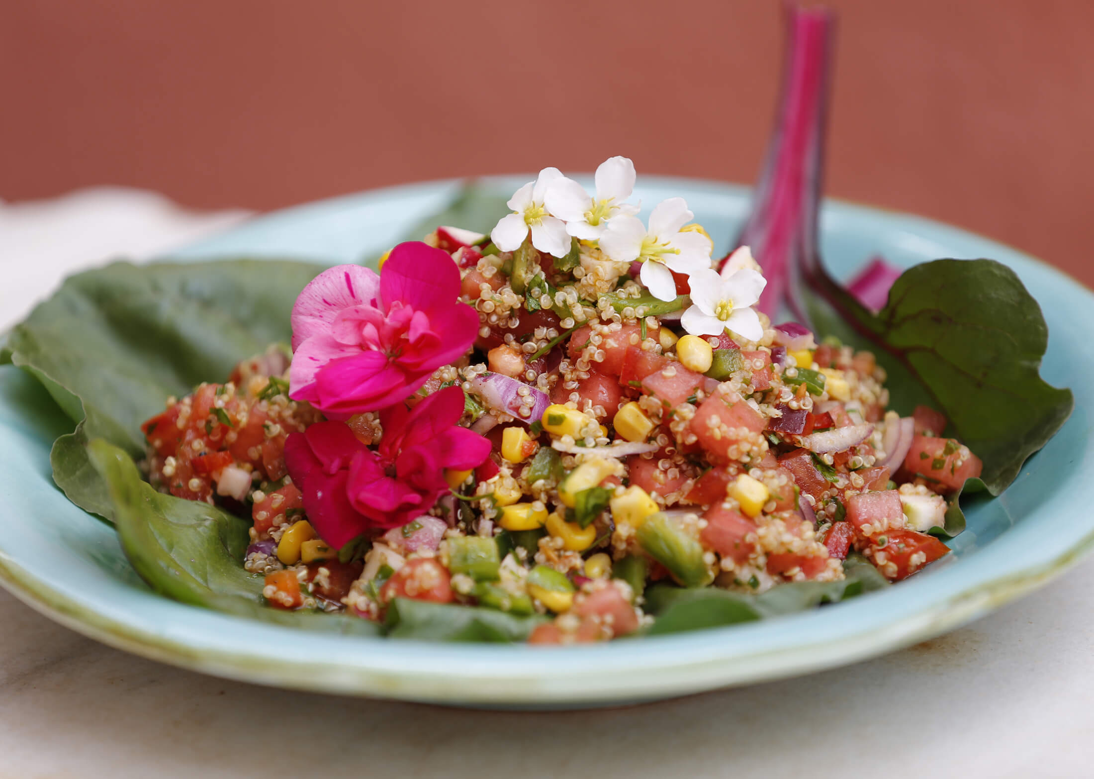 A colorful quinoa salad with diced vegetables and corn, garnished with pink and white edible flowers on a green leaf in a light blue bowl.
