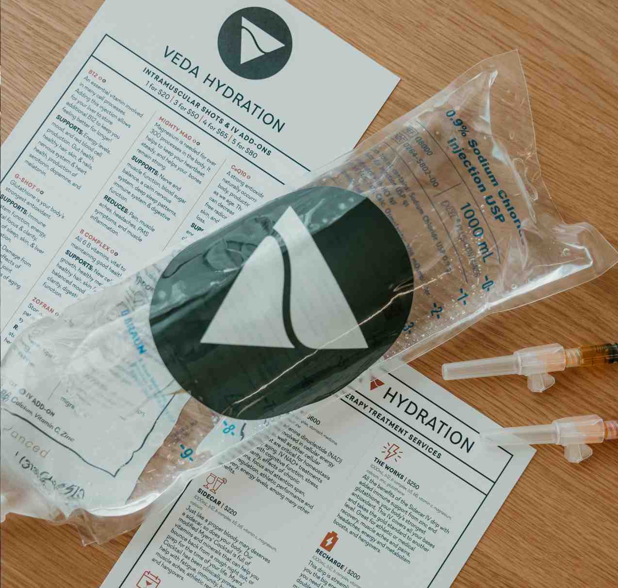 IV hydration saline bag and two syringes placed on top of printed hydration and therapy treatment service menus on a wooden surface.