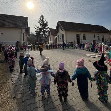 Óvodás gyerekek farsangi jelmezben körben állnak az udvaron, közös zenés foglalkozáson vesznek részt.
