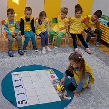 Egy óvodás kislány egy Bee-Bot tanulórobotot irányít egy számos és képes táblán, míg a többi gyermek figyelmesen követi a tevékenységet.