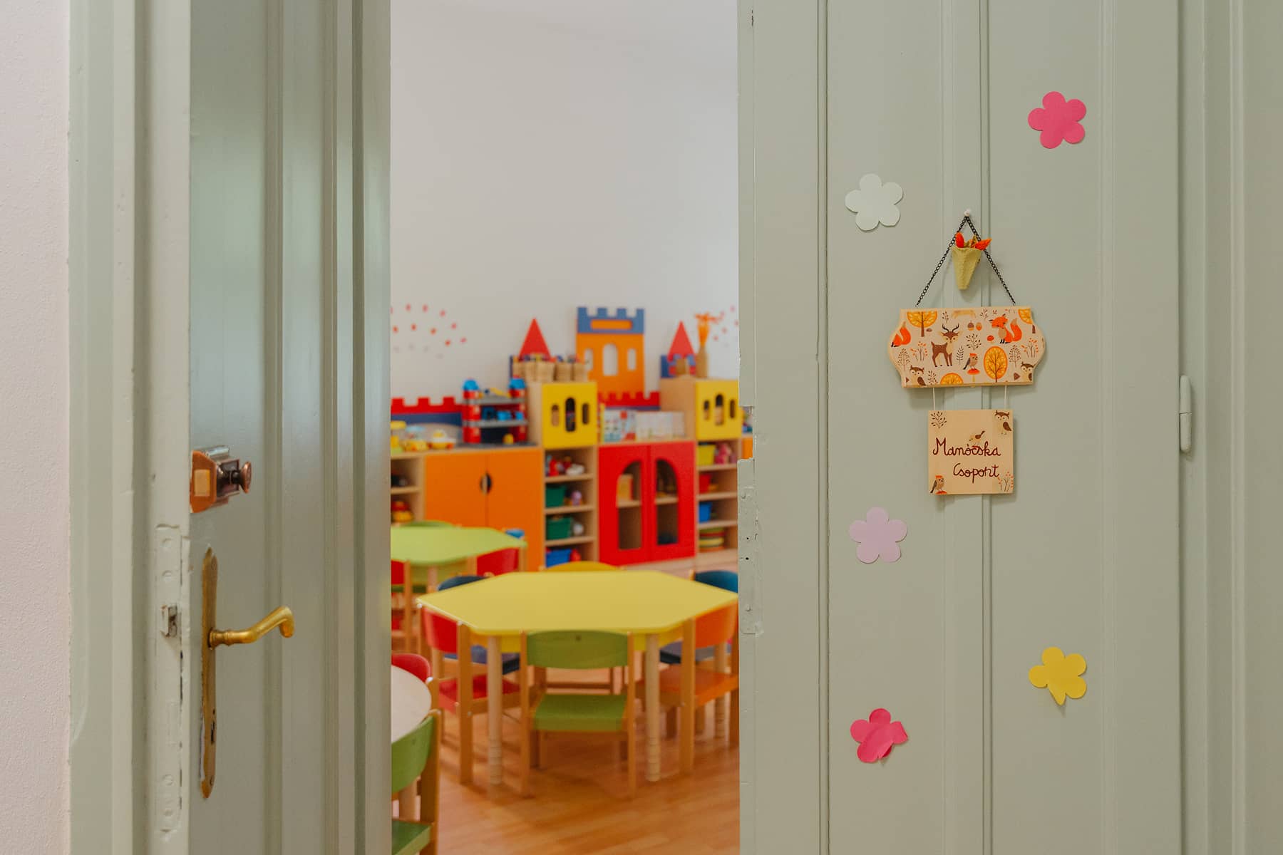 Óvodai csoportszoba nyitott ajtón keresztül fotózva, játékokkal és gyerekasztalokkal berendezve.