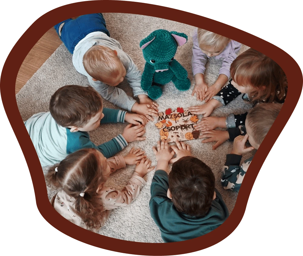 A Mazsola csoport gyermekei körben ülnek egy szőnyegen, középen egy Mazsola plüssfigura és a csoport neve látható