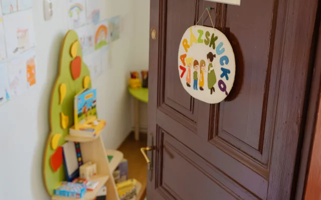 A Varázskuckó nyitott ajtaja, rajta varrott Varázskuckó feliratú tábla, a háttérben polcokon játékok és gyermekrajzok láthatók.