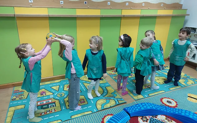 Az óvodások sorban állva mozgásfejlesztő labdajátékot játszanak, színes játszószőnyegeken a csoportszobában.