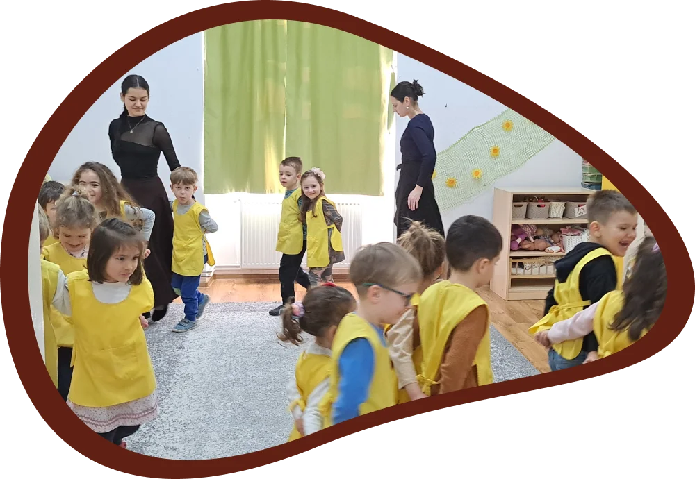 Csoportszobai néptánc foglalkozás, ahol a gyerekek követik az oktatók irányítását.