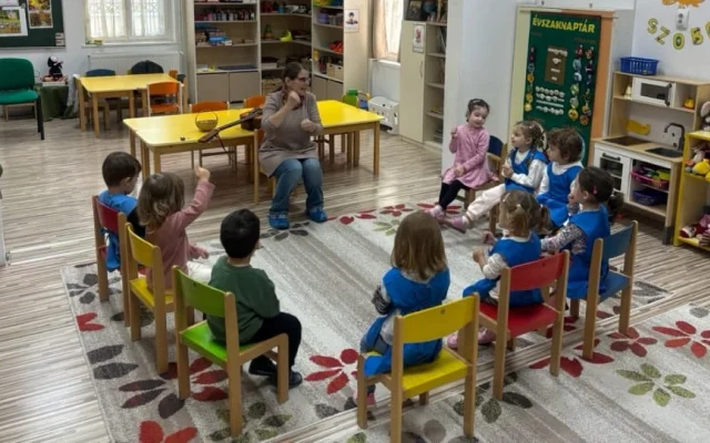 Az óvodás gyermekek körben ülve énekelnek és ritmusjátékot játszanak Kingrica vezetésével a csoportszobában.