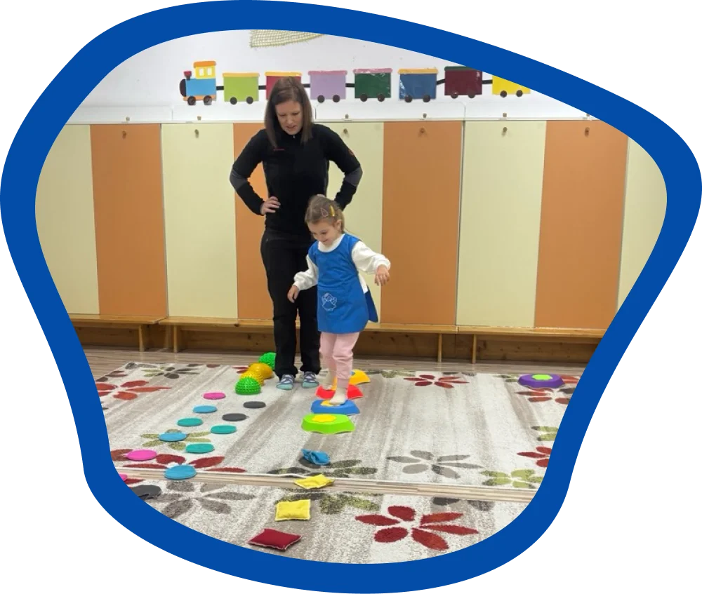 Óvodai csoportszobában egy kislány mozgásfejlesztő eszközökön halad át, miközben a pedagógus támogatja és figyelemmel kíséri a feladat végrehajtását.