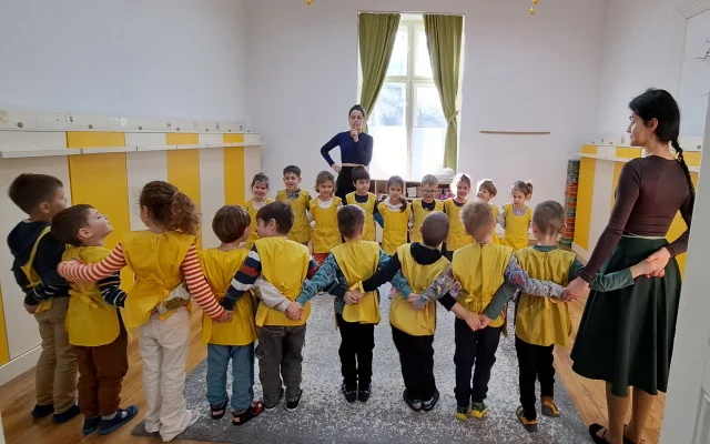 A néptánc foglalkozáson a gyermekek két sorban, egymás vállát átölelve állnak, és figyelik az oktatók instrukcióit a következő táncmozdulatokra vonatkozóan.