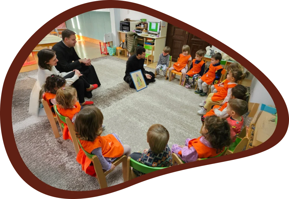 A csoportszobában színes székeken ülő óvodások figyelnek, miközben két plébános és a hitoktató vallási témájú képet mutat be és beszélget velük a foglalkozás során.