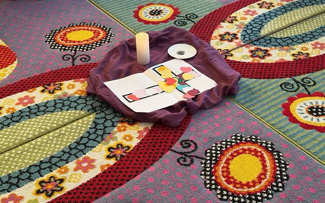 Színes, mintás óvodai szőnyegen, lila kendő, amelyre egy gyertya és színes papír szívecskékkel díszített nyomtatott kereszt van ráhelyezve.
