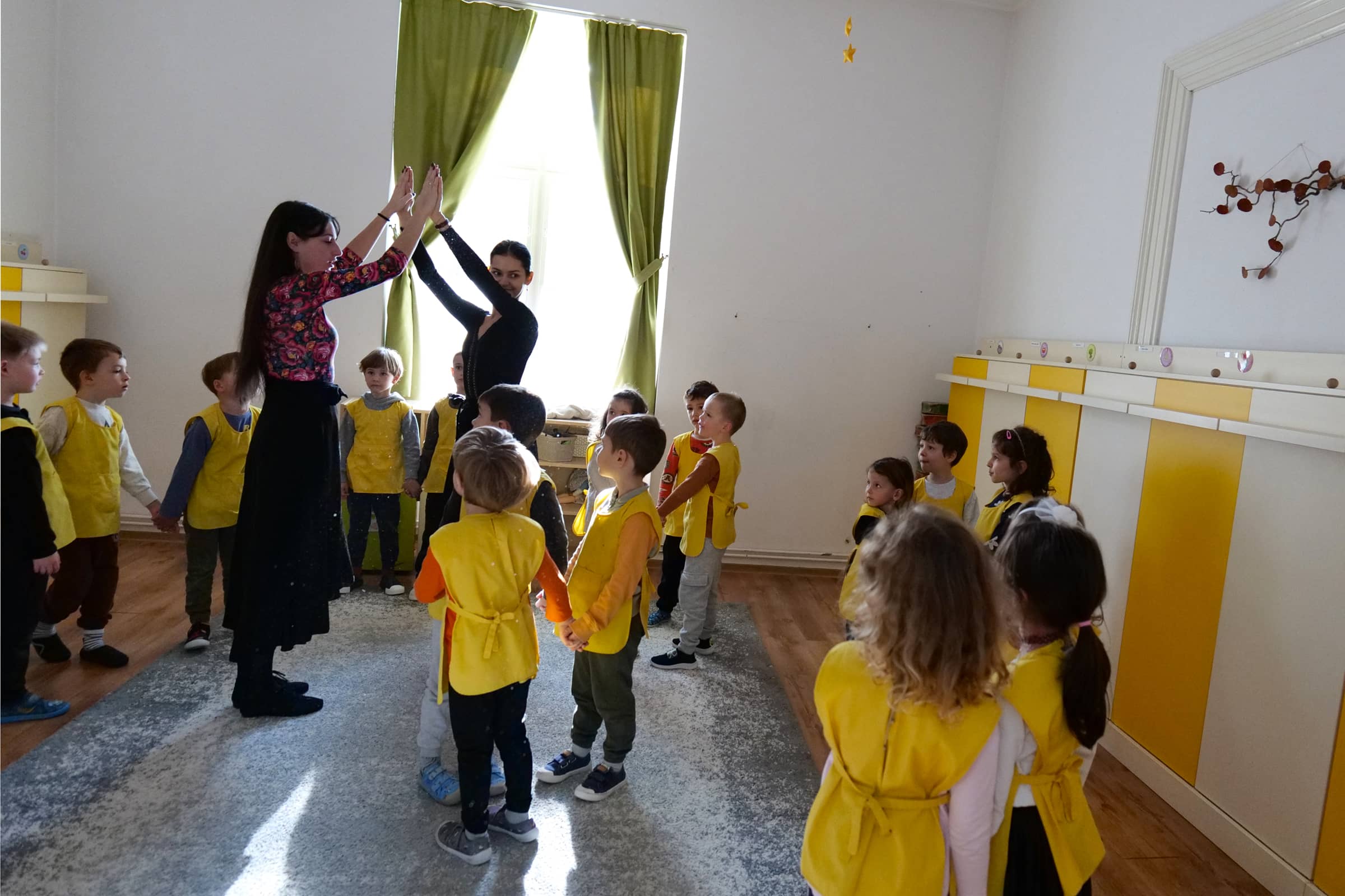 Ének- és táncfoglalkozás óvodában: a gyermekek körben figyelik a pedagógusok mozdulatait.
