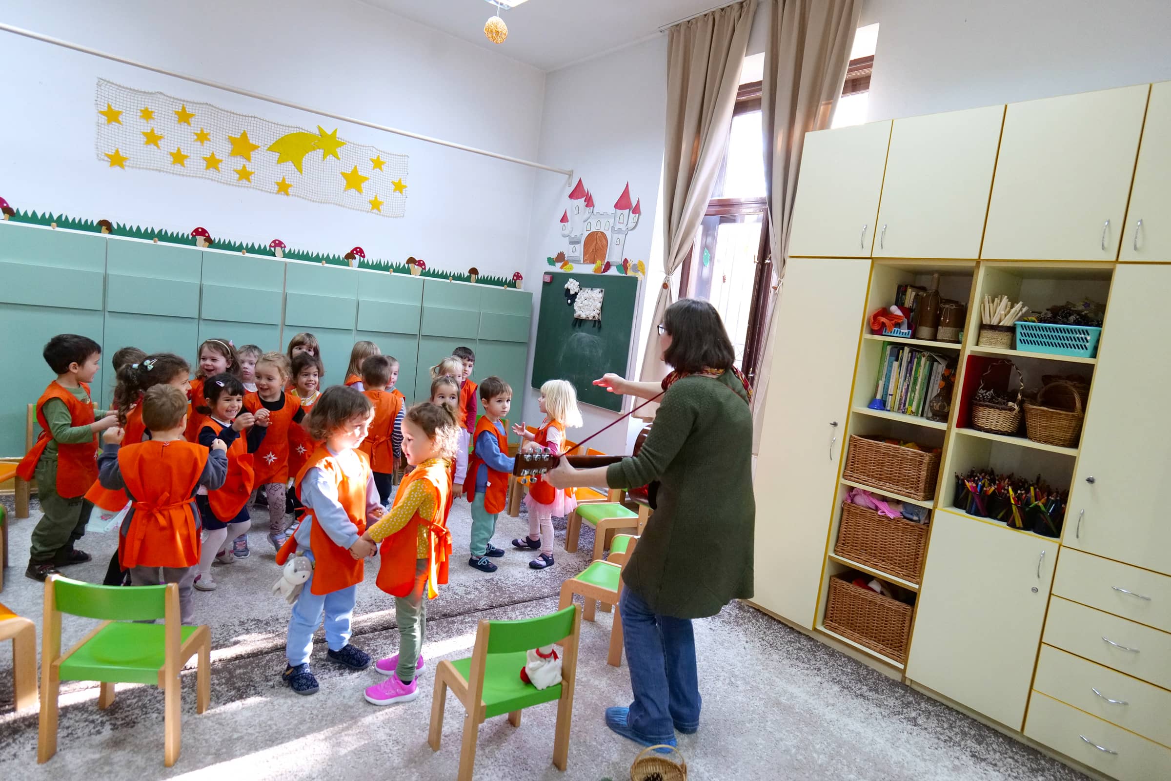 Óvodások közösen énekelnek és táncolnak a teremben, miközben a pedagógus gitáron kíséri őket.